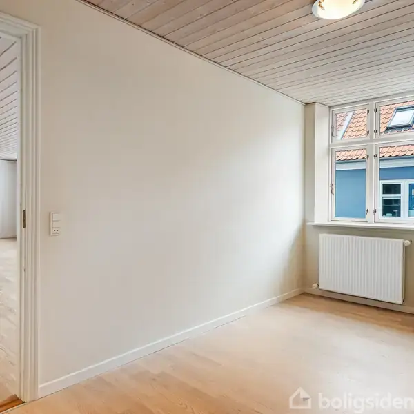 Et lyst rum med trægulv og hvidmalede vægge har en radiator under et vindue med udsigt til hustage. En tilstødende stue kan ses gennem en døråbning.
