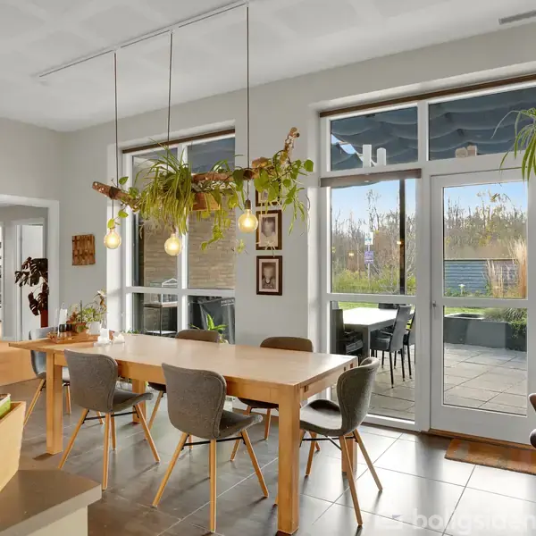 Et rektangulært spisebord står i en spisestue, omringet af seks stole. Rummet er lyst med store vinduer og flere hængende planter, der skaber en hyggelig atmosfære.