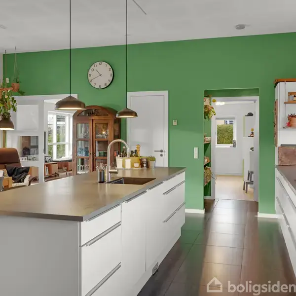 Køkken med hvid ø og indbygget vask, tre hængende lamper; grønmalet væg med ur, spisebord med stole; stue i baggrunden. Planter hænger fra loftet.