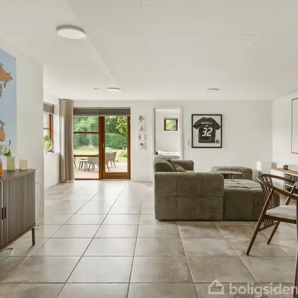 Stue med gråt sofaarrangement og spisebord; kort på væggen, sportsbluse i ramme. Store vinduer åbner ud til have med terrasse. Keramikfliser på gulvet.