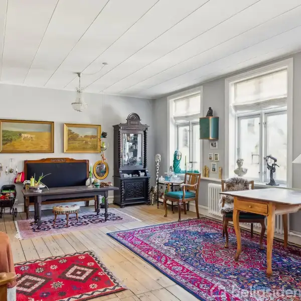 Stue med antikke møbler domineret af klaver og bord. Rummets træmøbler og tæpper skaber en klassisk atmosfære. Store vinduer lukker masser af naturligt lys ind.