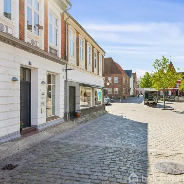En brostensgade med hvide og røde bygninger på venstre side. En åben plads strækker sig fremad med grønne træer og et busstoppested. En cykel står til højre.