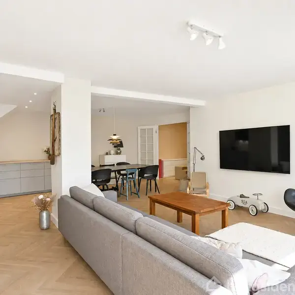 Grå sofa står i stue med fladskærm på væggen; hjørnekøkken til venstre med moderne indretning. Spisebord med stole i baggrunden, lys trægulv og hvidmalede vægge.