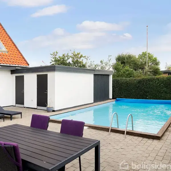 Svømmebassin med stige står stille ved siden af en terrasse med sorte liggestole og spisebord. Omkranset af et hvidt hus og grønne buske under en blå himmel.