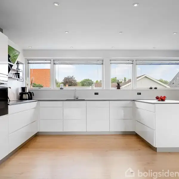 Hvidt køkken med moderne skabe står i et rum med trægulv og loftspots. Store vinduer i baggrunden giver udsigt til hustage og himmel. Assistenter står på bordpladen.