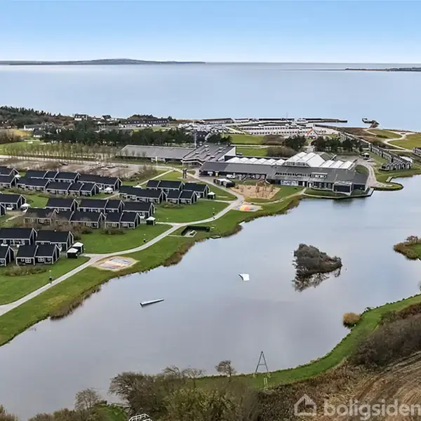 Bebyggelse med rækkehuse omgiver en sø; både ligger stille. I baggrunden ses havn og hav, flankeret af skove og enge.