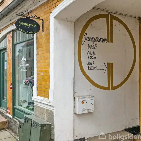 By med røde hustage langs en kystlinje. En pil peger mod et specifikt hus, mens en havn med både ses i baggrunden. Omgivet af grønt landskab.