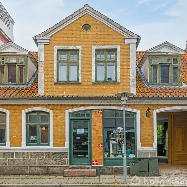 To-etagers hus med gul murstensfacade og rødt tegltag, har grønne vinduer og en træport. En butik ses i stueetagen med et skilt og et dansk flag ved vinduet. Gade som kontekst.