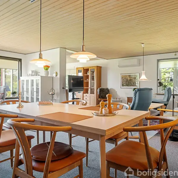 Spisebord med træstole står i en stue. Rummet har træloft, hængelamper og store vinduer, der lader lys komme ind. Der er også sofaer og et tv i baggrunden.