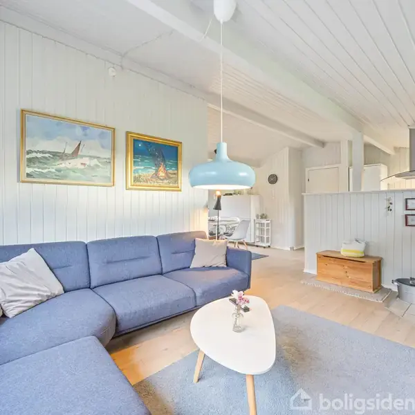 En blå hjørnesofa med puder står i en stue. Over sofaen hænger to malerier på væggen. En brændeovn er tændt til højre, og en lyseblå lampe hænger over et lille sofabord.