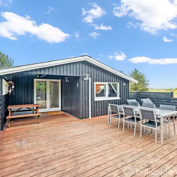 Et sommerhus med sort træfacade og store vinduer står på en træterrasse. Der er et bord-bænkesæt og et spisebord med stole. Haven er omgivet af grønne træer og græs.
