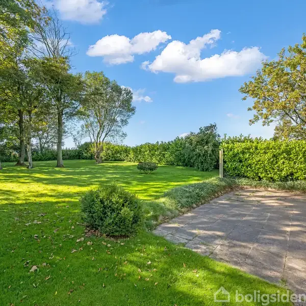 Terrassen ligger ved et hus, omgivet af en frodig grøn have med buske og træer under en blå himmel med skyer. En stol står på terrassen.