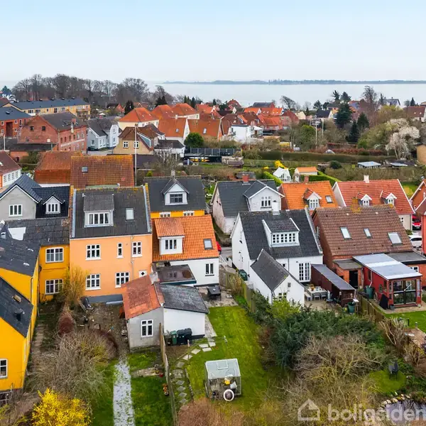 Flere farverige huse står tæt sammen i række. De har haver og små skure, med en by og havudsigt i baggrunden. Vandet kan ses i horisonten.