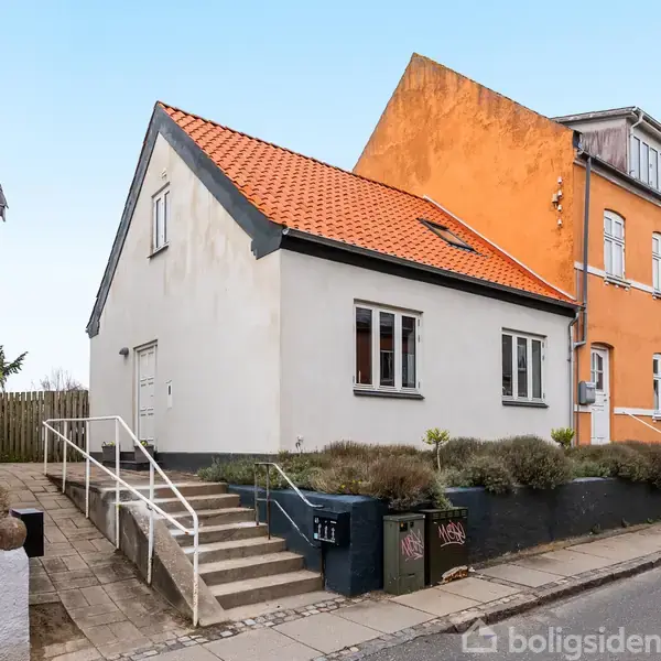 Hvidt hus med rødt tag står ved en fortovsgade. Trapper fører til hoveddøren, omgivet af små buske. Nabohuset har en skarp orange facade. Flisebelagt fortov og gaderække ses.