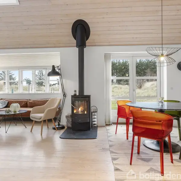 Brændeovn står midt i en lys stue og opvarmer rummet. Til venstre er en sofa med stole, og til højre er et spisebord med farverige stole foran vinduer.