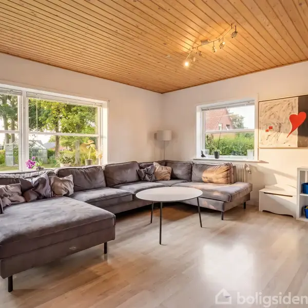 Grå hjørnesofa står ved to store vinduer med udsigt til haven. Rundt sofabord er foran sofaen. Væglampe og farverigt maleri med rød hjerteform i baggrunden.