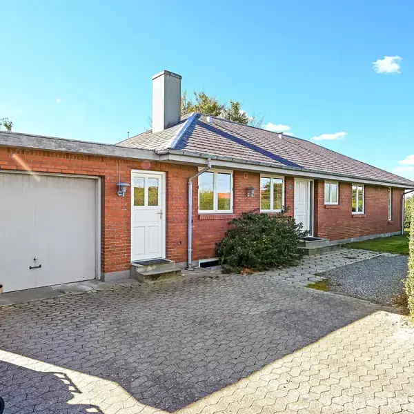 Rødt murstenshus med garage til venstre, hvid hoveddør og vinduer foran. En flisebelagt indkørsel fører op til huset, omgivet af buske. Klar blå himmel i baggrunden.