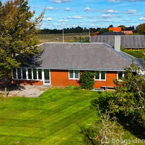 Et murstenshus med gråt tag står stille omgivet af en velholdt græsplæne og store træer. I baggrunden ses en åben mark og flere boliger.