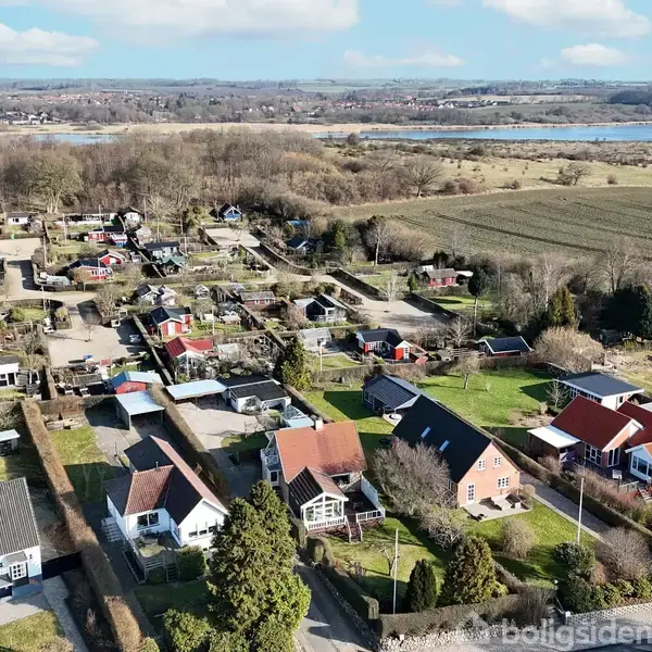 En række huse står stille i et parcelhuskvarter omgivet af marker og skove, med en flod i baggrunden. Solen skinner, og omgivelserne er landlige og fredelige.