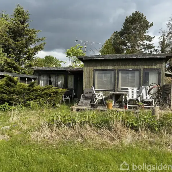 Et træhus står stille i en frodig have omgivet af høje træer og buske. Foran huset er der en terrasse med patio-møbler, der giver en rustik atmosfære.