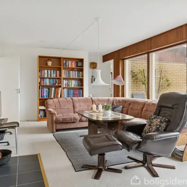 Brunt læderlænestol og sofa står i en stue med gul murstenspejs. Et sofabord er centralt placeret. Store vinduer giver udsigt til det grønne udenfor. Bogreol og fladskærms-TV er synlige.