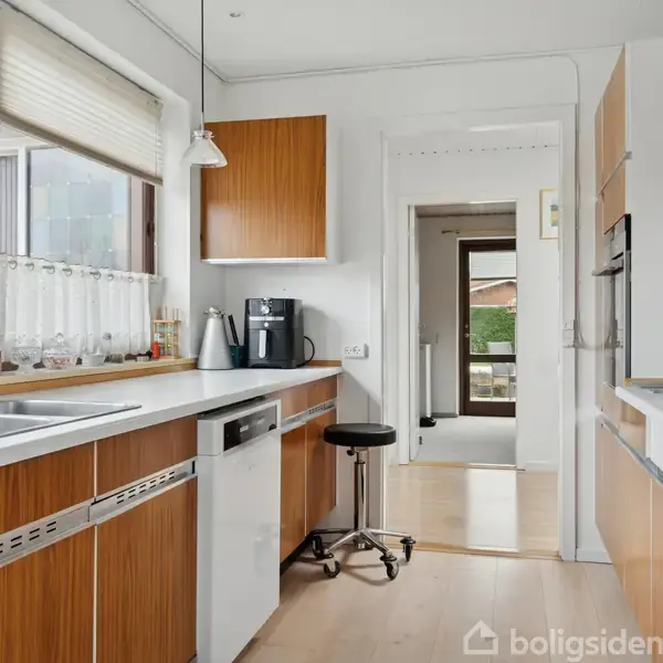 Et køkken med træskabslåger og hvid bordplade. En kaffemaskine står ved siden af en taburet foran et vindue med hvide gardiner. En dør leder til en lys gang.