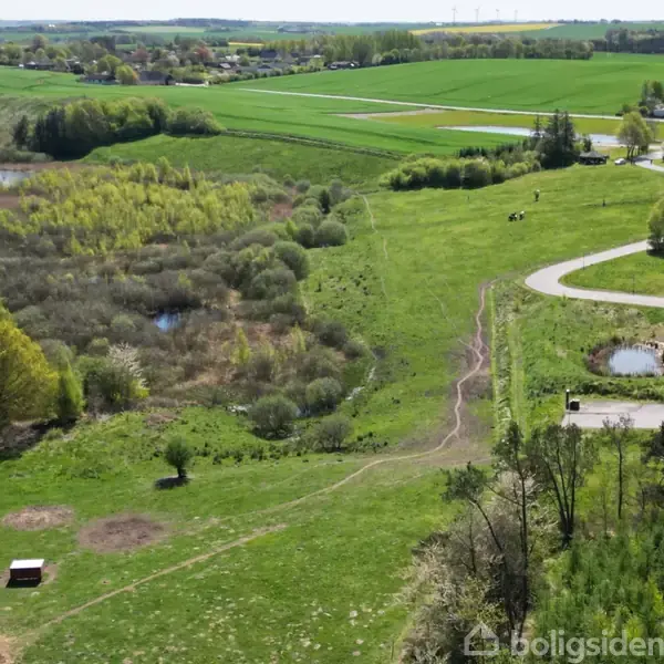 Grønt landskab set fra oven, med åbne marker, små søer og skovområder. Stier snor sig gennem terrænet nær en vej, mens en by kan anes i baggrunden.