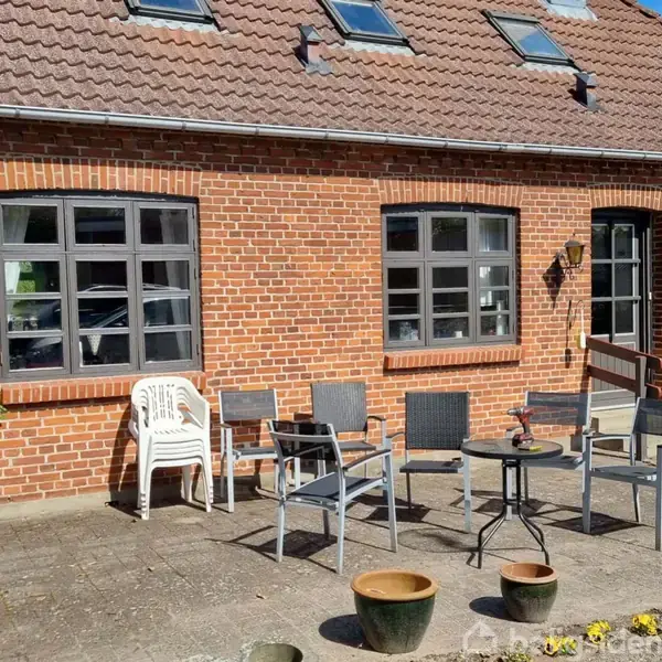 Stole og borde står på en flisebelagt terrasse foran en rød murstensbygning med sorte vinduer og døre. Ovenpå er der flere ovenlysvinduer. Forskellige potteplanter står i forgrunden.