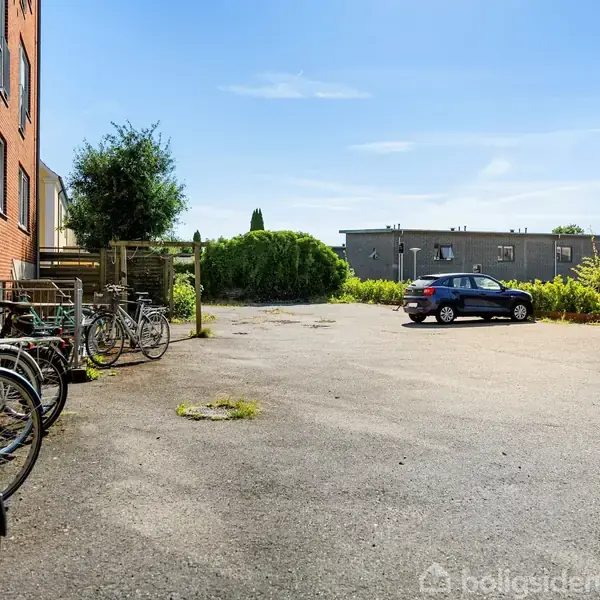 Cykler står parkeret mod en rød murstensbygning, mens en sort bil holder parkeret på en åben asfaltplads. Grønne buske og træer i baggrunden under en klar himmel.