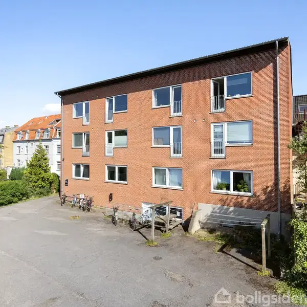 Bygning med røde mursten står stille i sollys, omgivet af en asfalteret gårdsplads og cykler, med grønne buske og træer i baggrunden.