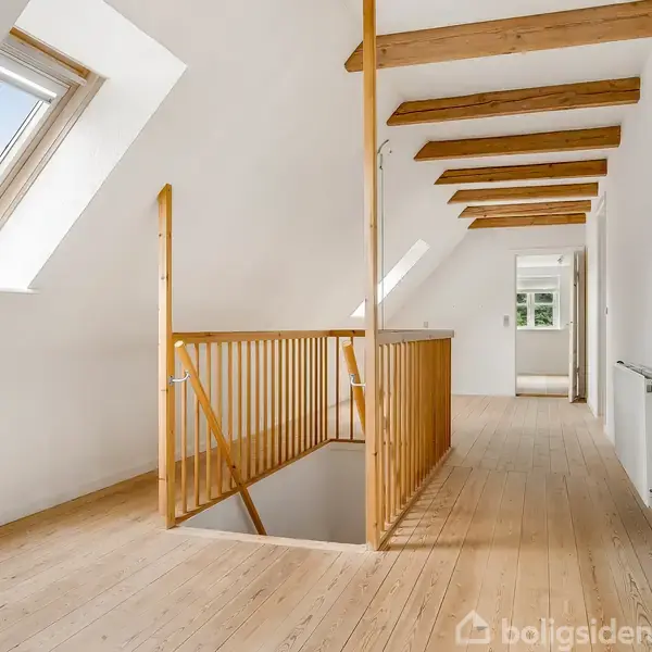 Trætrappe fører op til en lys hems med træbjælker i loftet, hvidmalede vægge og et skråvindue, der giver naturligt lys. En radiator er placeret på højre væg.