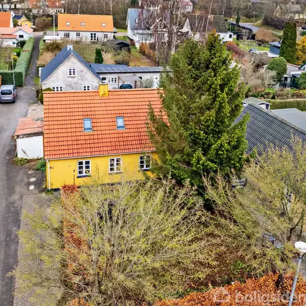Hus med gult facade og rødt tag står i forstadsområde omgivet af træer og andre boliger. En smal vej løber forbi huset, omgivet af flere andre bygninger og haver.