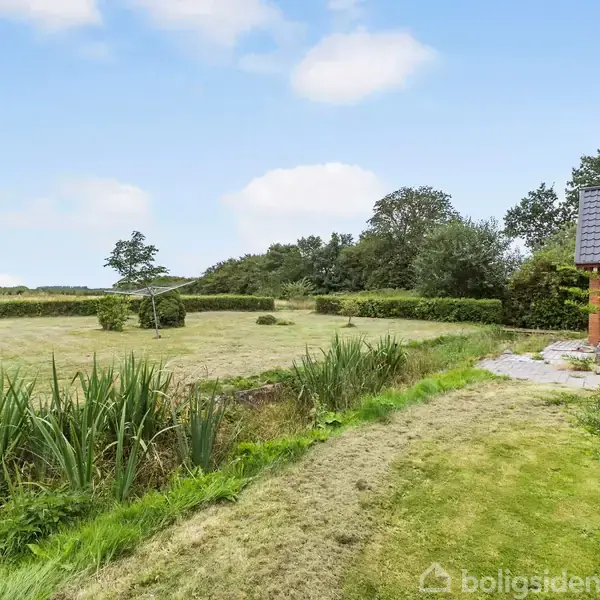En murstensbygning står ved en velplejet græsplæne, omgivet af buske og træer i landlige omgivelser. Himlen er blå med enkelte skyer.