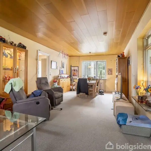 Stue med polstrede stole og sofa, dekoreret vitrineskab til venstre, fylderummeligt rum med træloft og tæppe, store vinduer i baggrunden og diverse indretningsgenstande.