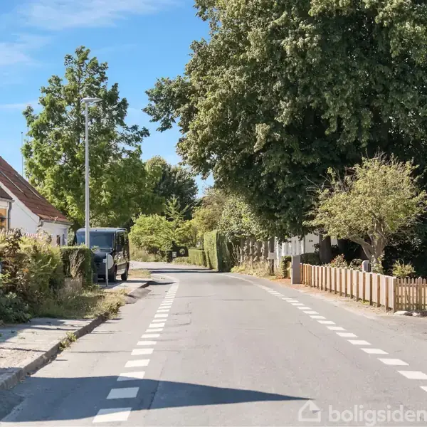 Vejen strækker sig gennem en landsby med huse og træer langs kanterne, mens en blå himmel ses i baggrunden.