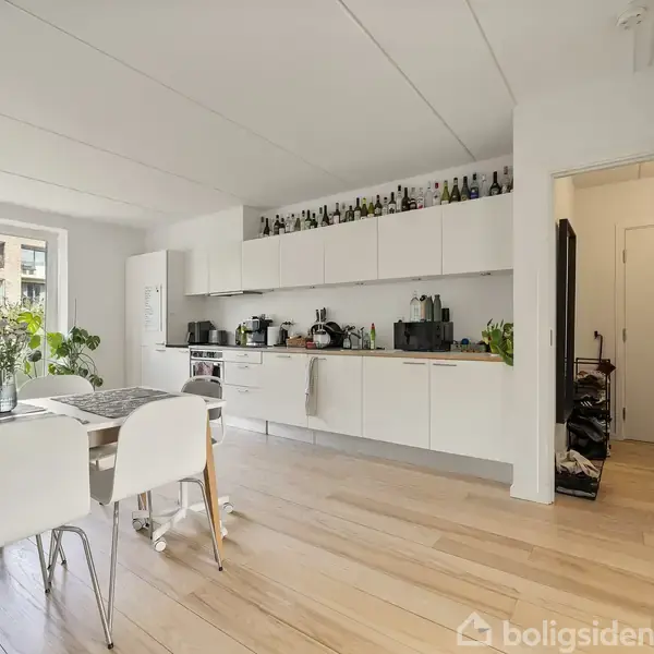 Et moderne køkken med hvide skabe og apparater på bordpladen. Et spisebord med fire stole står i forgrunden. Der er en døråbning til en gang i baggrunden.