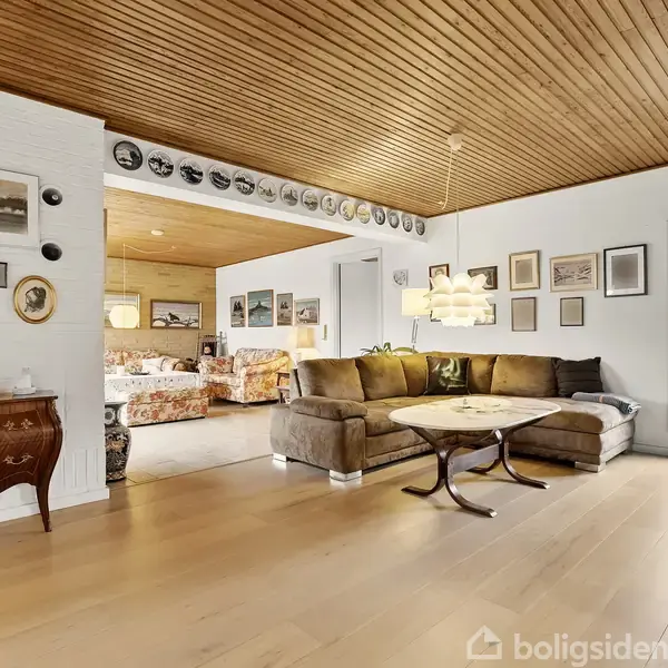 En stor, brun sofa står i et åbent rum med trægulv og træloft. Væggene er dekoreret med billeder og tallerkener. Et vindue giver udsigt til en have.