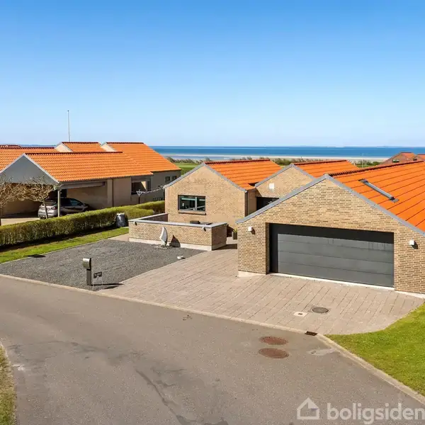 Hus med rødt tag står stille i et roligt nabolag med udsigt til havet i baggrunden. Flisebelagt indkørsel og garage foran huset.