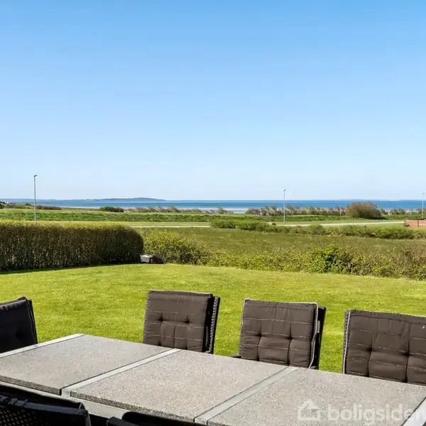En udendørs spisebord med stole står på en terrasse. Det har udsigt til en velplejet græsplæne og en bølgende kystlinje i det fjerne under en klar blå himmel.