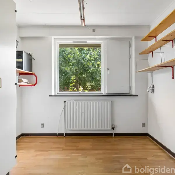 Et tomt rum med trægulv har en hvid væg og et vindue med udsigt til grønne træer. En radiator er placeret under vinduet, og der er hylder på højre væg.