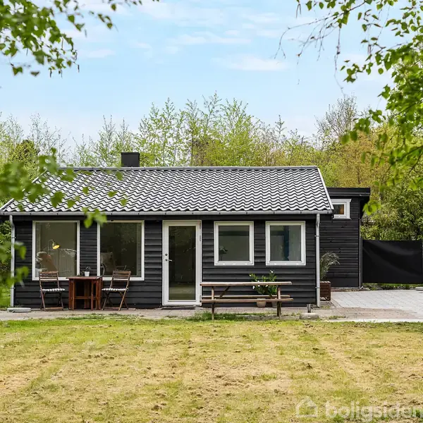 Et sort træhus med hvide vinduer står stille i en grøn have omkranset af træer. Der er et bord og stole på terrassen foran huset.