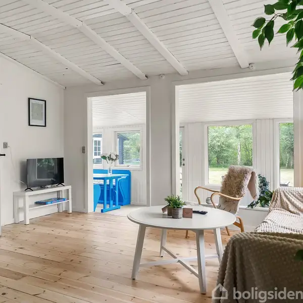 Stue med hvidmalet træloft og plankegulv, sofa og rundt bord med planter. Tv'et står på en reol imod væggen. I baggrunden ses et rum med blå stole og et spisebord.