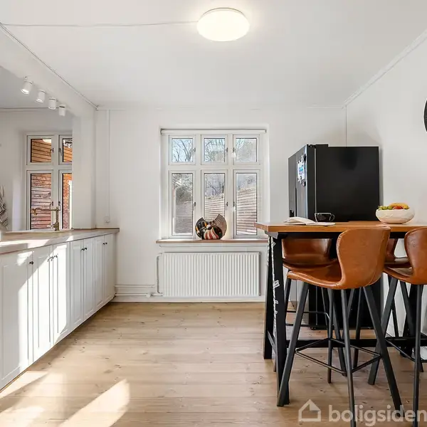 Et moderne køkken med en høj sort bordplade, omgivet af høje stole, en sort køleskab og en vægur på væggen. Lyse trægulve og et vindue i baggrunden.