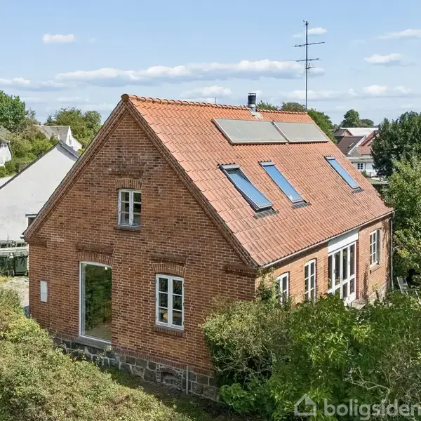 Rødt murstenshus med solpaneler på taget, vinduer på begge etager, og omgivet af grønne buske og træer. Beliggende ved en landevej med flere huse i baggrunden.