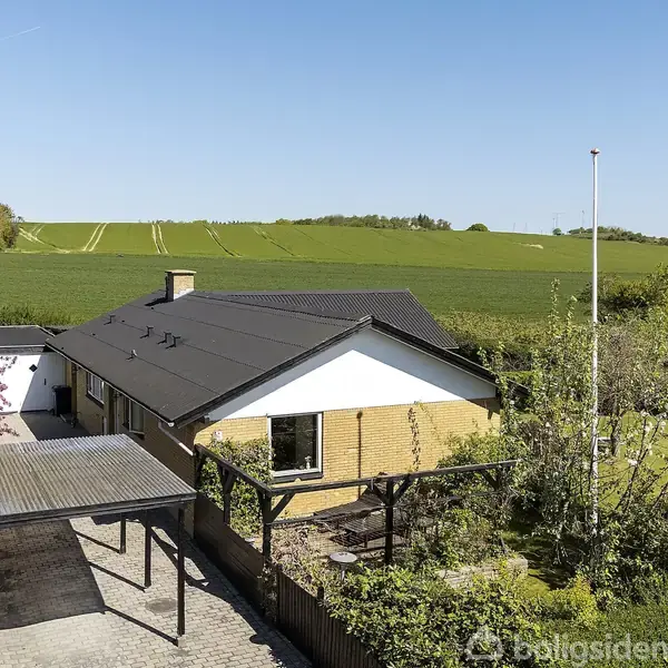 Et enfamiliehus med gult murværk har en sort tag og carport. Det er omgivet af en velholdt have og ligger nær åbne marker under en klar blå himmel.