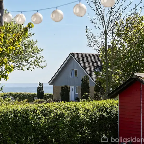 En grå villa med rødt tag ses blandt grønne træer. I forgrunden hænger lyskæder, og der er et rødt skur. I baggrunden ses havet og blå himmel.