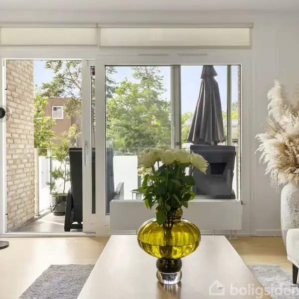 En stue med et sofabord i forgrunden, der har en gul vase med blomster. Skydedøre fører ud til en balkon med udsigt til et grønt område.