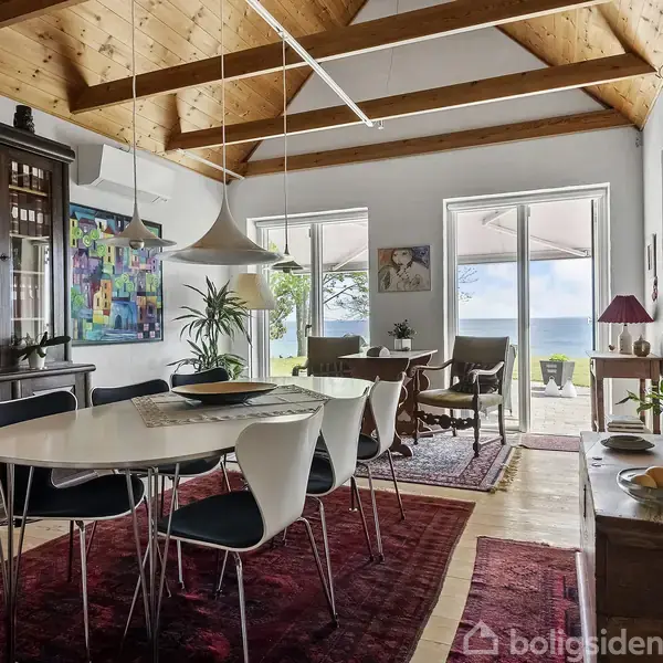 Spisebord med sorte stole står på et rødt tæppe, mens lys hænger fra loft med træbjælker. Store vinduer viser en terrasse med havudsigt. Ingen tekst synlig.