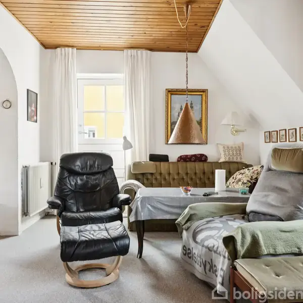 En stue med lænestole og en sofa placeret omkring et sofabord. Et vindue og kunstværker hænger på væggen. Rummet er lyst med hvide gardiner og en træbeklædt loft. Tekst på tæppe: "selsvtyke".