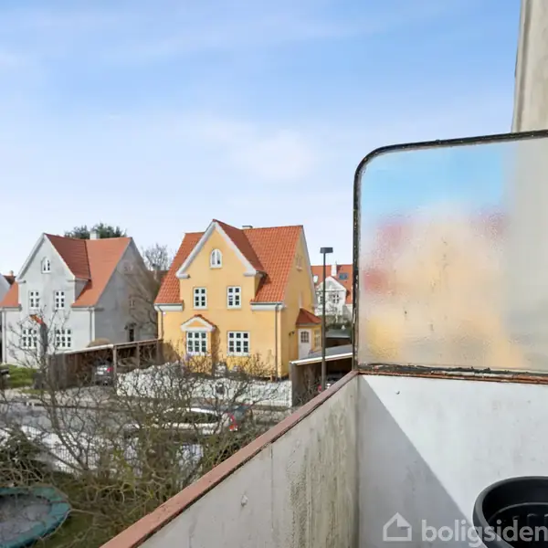 Balkon viser en udsigt over et boligområde med farverige, traditionelle huse og røde tage. En gennemsigtig skærm adskiller balkonen fra naboen. Træer og veje er synlige i baggrunden.